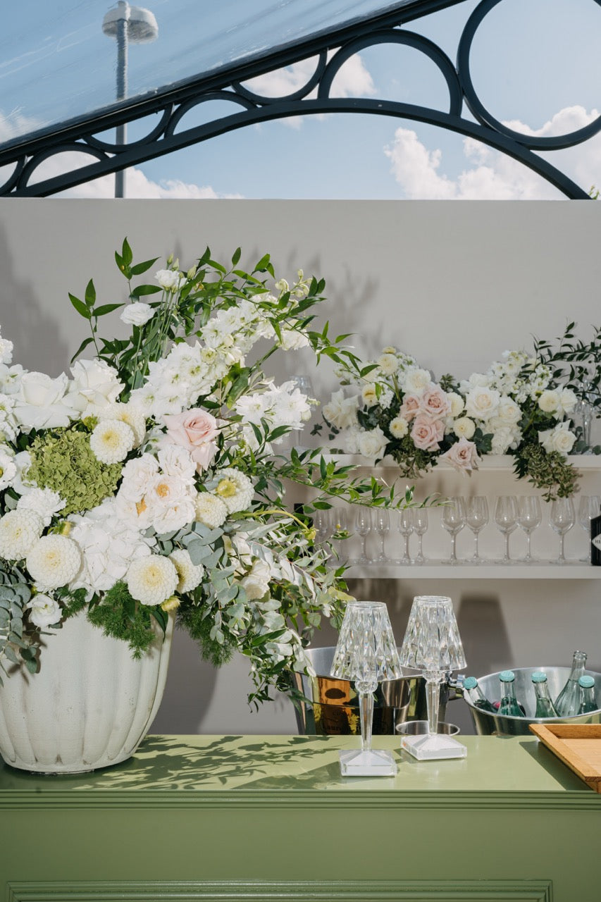 Stilvolles und ästhetisches Blumenarrangement mit weißen und rosafarbenen Blüten auf einer grünen Bartheke, umgeben von Kristalllampen deutschlandweit. Ein großes, elegantes Blumenarrangement aus weißen und rosafarbenen Blüten schmückt die grüne Bartheke. Kristalllampen und edle Details runden das luxuriöse Setting ab, das eine perfekte Mischung aus Eleganz und Natur bietet. Ideal für eine stilvolle Hochzeit im modernen Ambiente.
