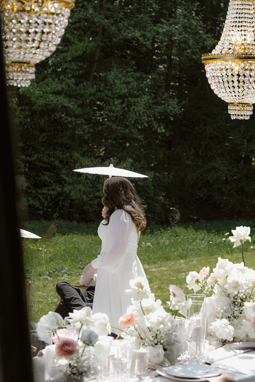 Braut im Garten mit weißem Papierschirm, umgeben von eleganter Hochzeitsdekoration und Kronleuchtern in Köln. Diese romantische Szene zeigt eine Braut, die im Garten mit einem weißen Papierschirm flaniert. Im Vordergrund sind edle Hochzeitsdekorationen mit weißen Blumen und funkelnden Kronleuchtern zu sehen, was eine stilvolle und verträumte Atmosphäre schafft.