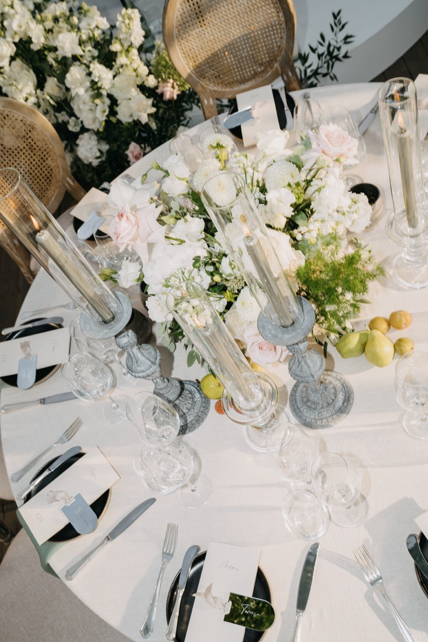 Dekorationskonzept mit einem elegantem Hochzeitstisch mit weißen Blumen, hohen Kerzenhaltern und stilvoll gedecktem Besteck. Dieses Dekorationskonzept zeigt einen Hochzeitstisch mit einer prächtige Blumendekoration aus weißen Blüten, umgeben von eleganten Kristall-Kerzenhaltern. Das stilvolle Besteck und die perfekt arrangierten Gläser unterstreichen die luxuriöse und romantische Atmosphäre, die für eine gehobene Hochzeit ideal ist.