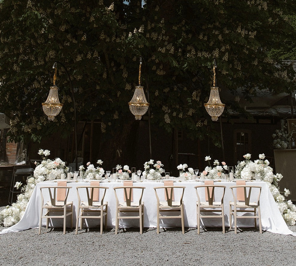 Edle Hochzeitsdekoration mit langem Tisch, weißen Blumenarrangements und hängenden Kronleuchtern im Freien. Diese romantische und elegante Hochzeitsdekoration zeigt einen langen, festlich gedeckten Tisch im Freien, geschmückt mit üppigen weißen Blumenarrangements und stilvollen, hängenden Kronleuchtern. Ideal für eine Outdoor-Hochzeit mit luxuriösem Flair.