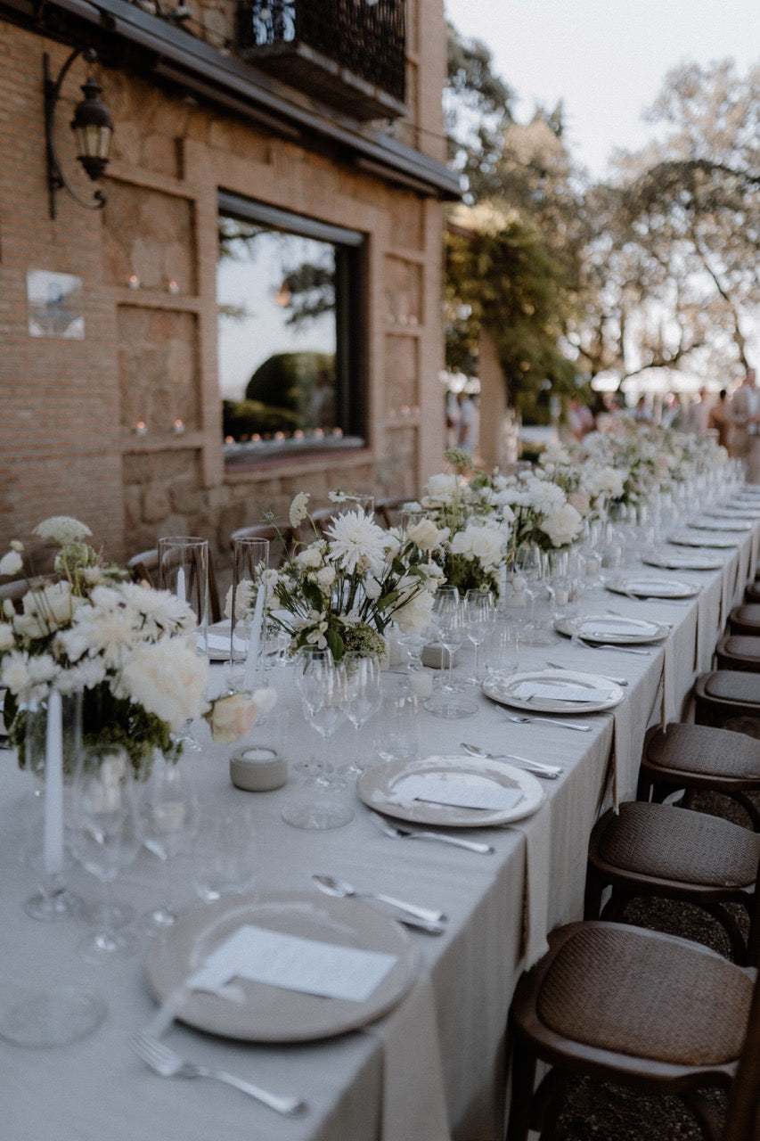 Elegante, ästhetische Hochzeitstafel im Freien mit weißen Blumenarrangements und rustikalen Holzstühlen. Eine stilvolle Hochzeitstafel im Freien vor einem steinernen Gebäude, dekoriert mit weißen Blumen, edlem Geschirr und rustikalen Holzstühlen, ideal für eine charmante und zeitlose Hochzeitsfeier.