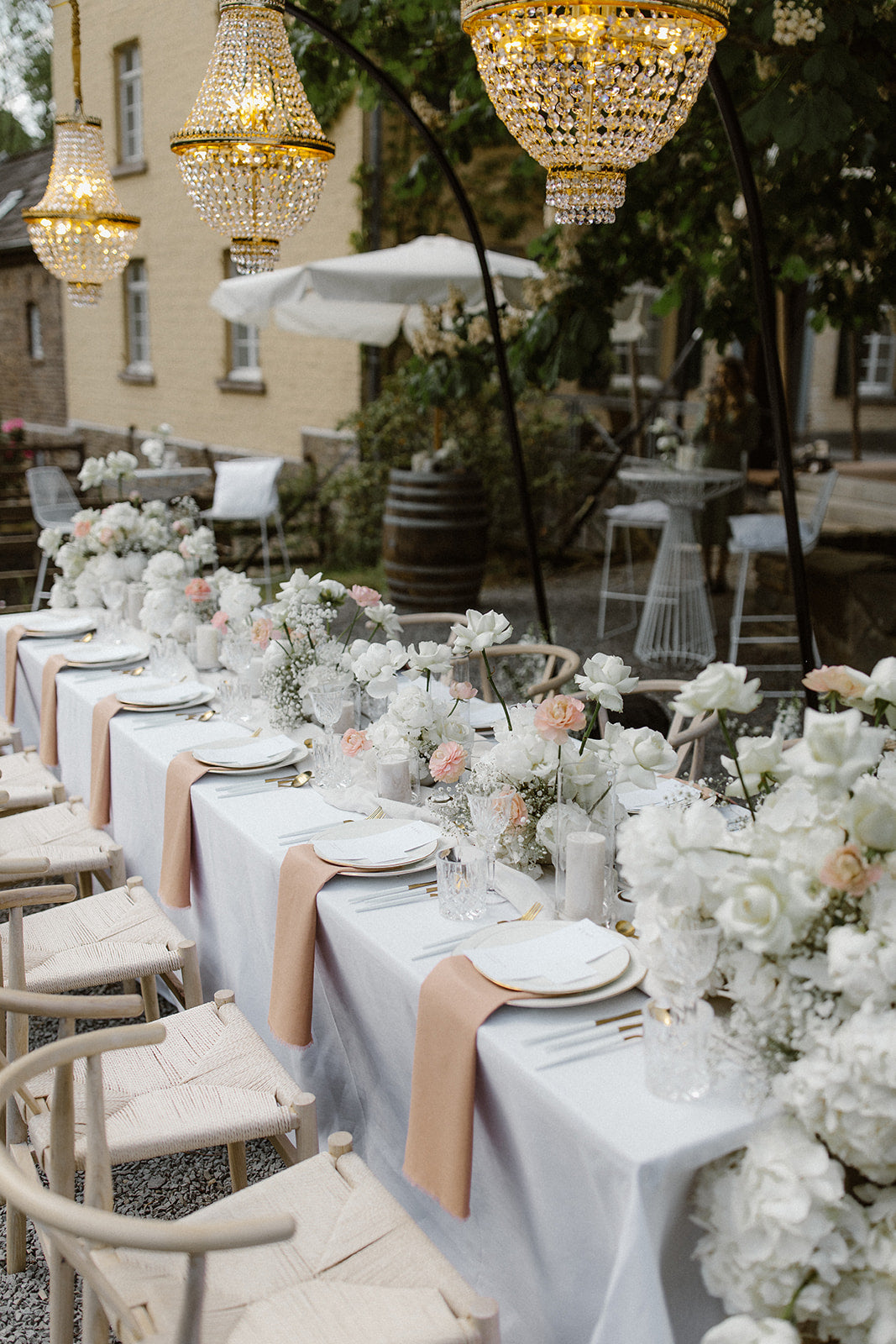 Lange Hochzeitstafel mit edler weißer Blumendekoration und hängenden Kronleuchtern im Freien. Diese elegante Hochzeitstafel im Freien ist mit weißen Blumenarrangements und luxuriösen hängenden Kronleuchtern dekoriert. Die harmonische Kombination aus natürlichen und glamourösen Elementen schafft eine stilvolle Atmosphäre für eine romantische Feier unter freiem Himmel.