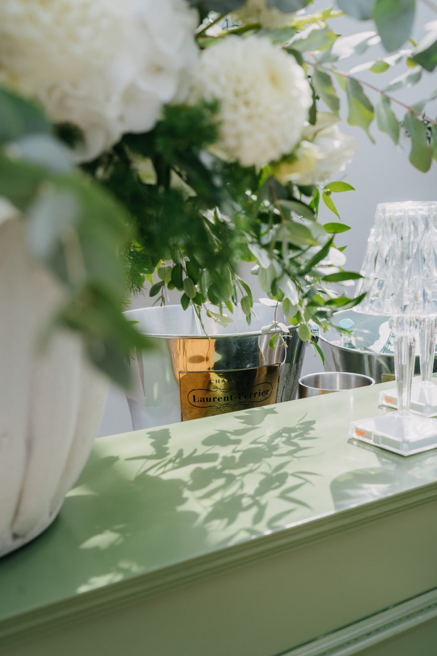 Detail einer Bar mit eleganter Blumendekoration und Kristallgläsern auf einer grünen Theke. Dieses Bild zeigt ein stilvolles Detail einer Hochzeitsbar, bei dem die üppige Blumendekoration in einer eleganten Vase und funkelnde Kristallgläser auf einer grünen Theke arrangiert sind. Das Zusammenspiel von natürlichen und luxuriösen Elementen schafft eine einladende und raffinierte Atmosphäre für die Gäste.