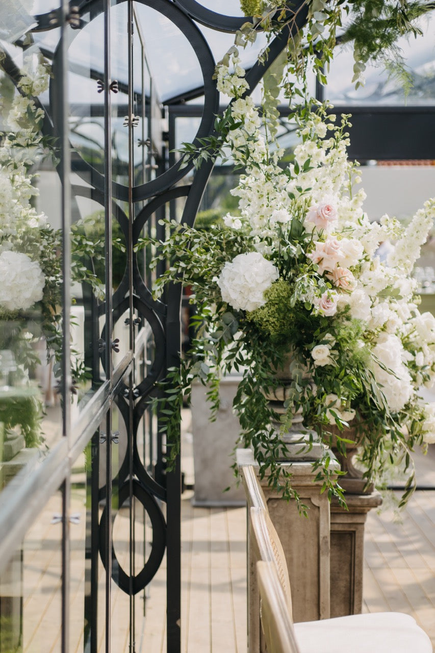 Ein prächtiges Blumenarrangement mit weißen und rosafarbenen Blüten in einer Orangerie in Berlin. Dieses große Blumenarrangement aus weißen und rosafarbenen Blüten, kombiniert mit üppigem Grün, schmückt stilvoll die Seiten der Orangerie. Die reflektierende Glasstruktur und die floralen Details schaffen eine harmonische Verbindung zwischen Natur und moderner Eleganz. Ideal für eine exklusive Hochzeit mit romantischer Atmosphäre.