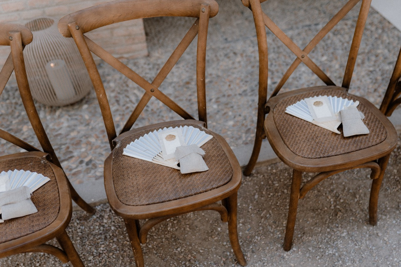 Schlicht dekorierte Hochzeitsstühle aus Holz mit Fächern und Servietten. Rustikale Holzstühle bei einer Hochzeit im Freien, dekoriert mit schlichten Fächern und Servietten, passend für eine minimalistische und charmante Feier.
