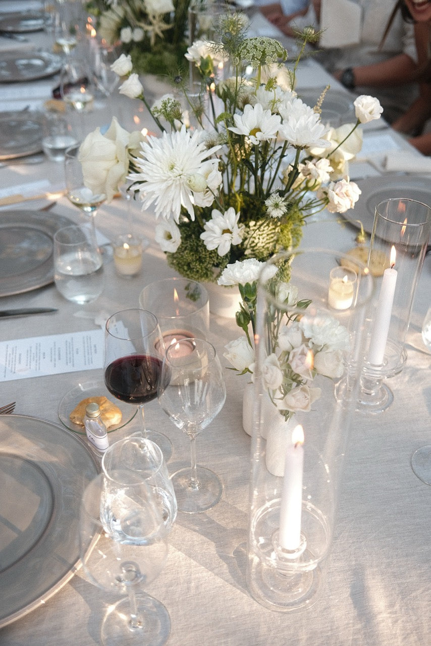 Hochzeitstafel mit weißem Blumenarrangement, Kerzen und elegantem Geschirr. Schlichte und elegante Dekoration einer Hochzeitstafel mit weißen Blumen, Kerzen und edlem Geschirr, perfekt für eine zeitlose und charmante Hochzeitsfeier.