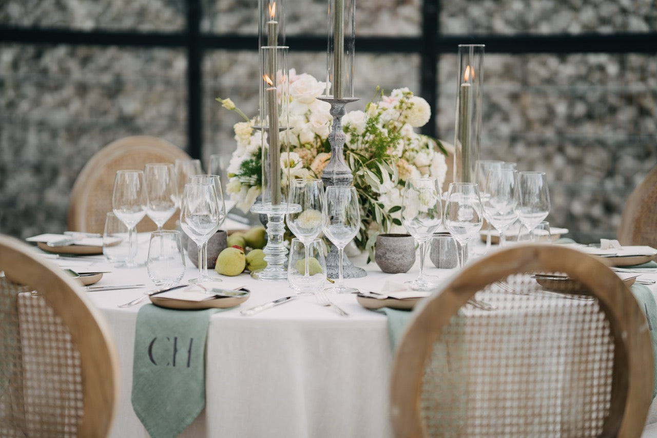 Hochzeitsinspiration eines stilvoll gedeckten Hochzeitstisches mit Blumendekoration, hohen Kerzen und personalisierten Leinenservietten. Dieser elegant dekorierte Hochzeitstisch besticht durch ein zentrales Blumenarrangement aus weißen und grünen Blüten, ergänzt durch hohe Kerzen in Kristallhaltern. Die personalisierten Leinenservietten und die perfekt arrangierten Gläser und Teller verleihen dem Setting eine luxuriöse und individuelle Note. Ideal für eine Hochzeit, die auf Details und Eleganz setzt.