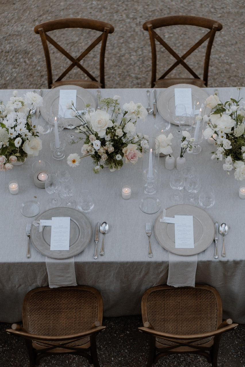 Schlicht gedeckter Hochzeitstisch mit weißen Blumenarrangements und rustikalen Holzstühlen. Eleganter Hochzeitstisch im Freien mit weißen Blumenarrangements, dezenten Kerzen und rustikalen Holzstühlen, ideal für eine minimalistische und zeitlose Feier.