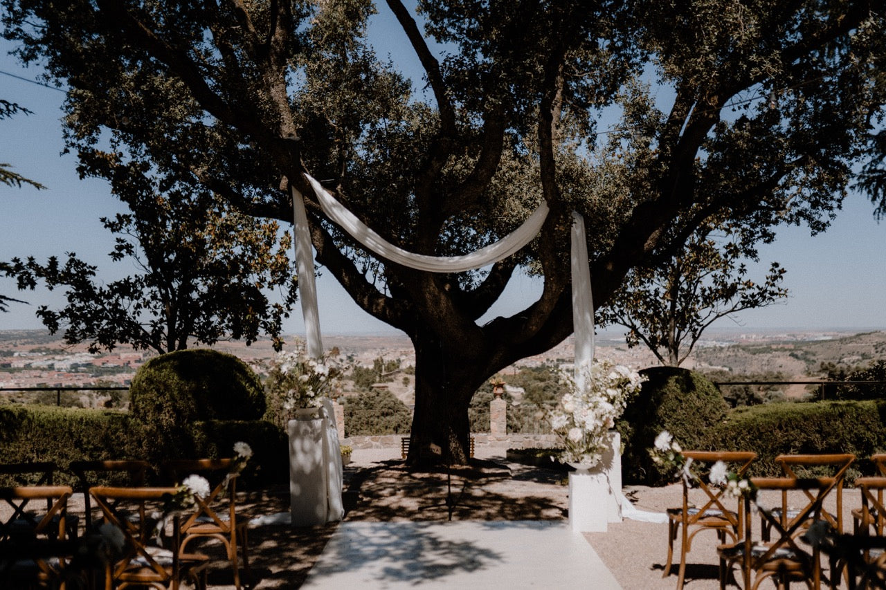 Schlicht dekorierte Trauung im Freien unter einem großen Baum mit weißen Stoffen und Blumen. Eine romantische Hochzeitszeremonie im Freien, mit einem eleganten Altar unter einem großen Baum, dekoriert mit weißen Stoffbahnen und Blumen, ideal für eine natürliche und zeitlose Feier.