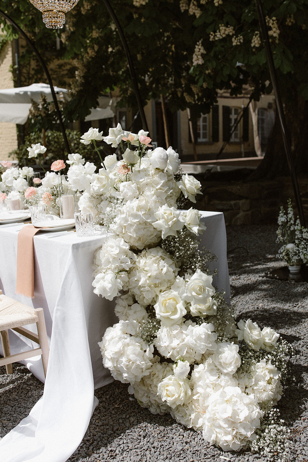 Ästhetischer Hochzeitstisch mit üppiger Blumengirlande aus weißen Blüten und Schleierkraut. Diese luxuriöse Hochzeitsdekoration besticht durch eine üppige Girlande aus weißen Blüten, die über die Seite des Tisches fließt. Das edle Blumenarrangement verleiht dem Tisch ein elegantes und romantisches Flair, perfekt für eine stilvolle Outdoor-Hochzeit.