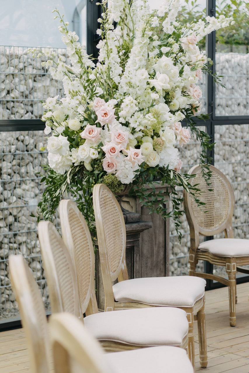 Eleganter Sitzbereich für die Trauung mit weißen Polsterstühlen und großem Blumenarrangement aus weißen und rosafarbenen Blüten bei einer luxuriösen Hochzeit. Der Trau-Sitzbereich dieser luxuriösen Hochzeit ist mit eleganten, weißen Polsterstühlen und einem üppigen Blumenarrangement aus weißen und rosafarbenen Blüten dekoriert. Die geschmackvolle Kombination aus hochwertigen Möbeln und floraler Pracht schafft eine festliche und exklusive Atmosphäre für die Hochzeitszeremonie. Ideal für Paare, die auf Detail