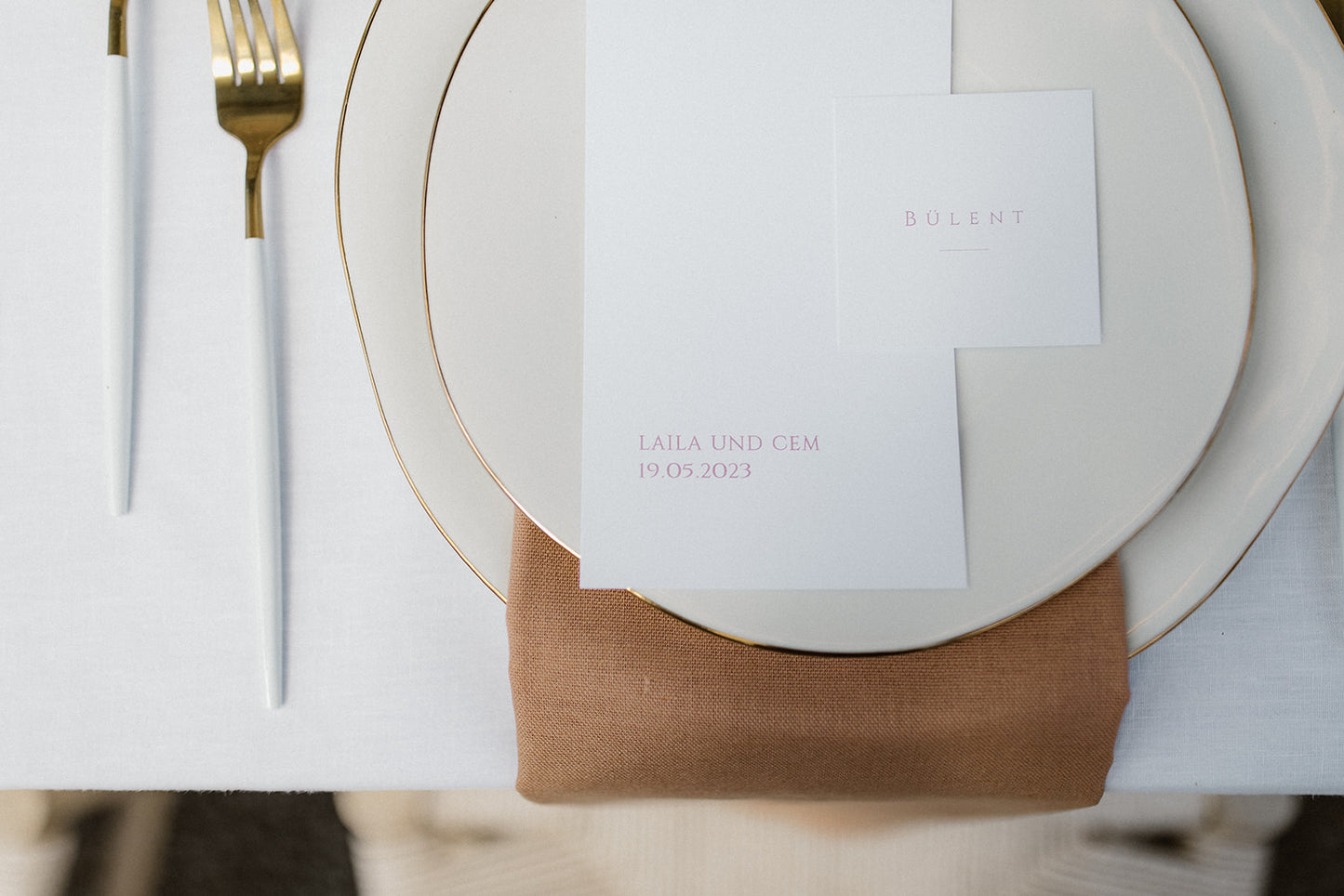 Minimalistisches Hochzeitstischgedeck mit Menükarte und goldenen Besteckteilen. Dieses minimalistische Hochzeitstischgedeck besticht durch eine schlichte Menükarte auf cremefarbenem Geschirr, ergänzt durch edle goldene Besteckteile und eine erdige Stoffserviette. Die dezente Eleganz verleiht dem Tisch ein modernes und stilvolles Ambiente.