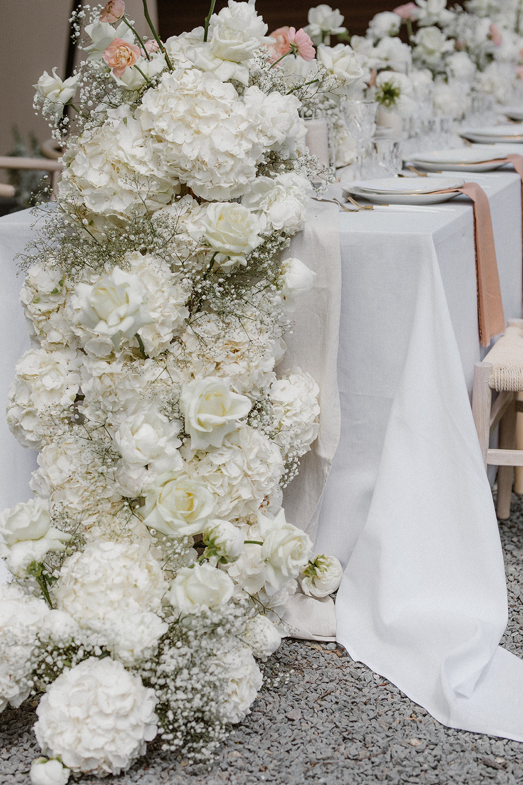 Üppige weiße Blumendekoration aus Hortensien entlang eines Hochzeitstisches. Diese opulente Hochzeitsdekoration besticht durch eine üppige Blumenarrangement aus weißen Hortensien und Schleierkraut, das entlang eines Hochzeitstisches drapiert ist. Perfekt für eine elegante und romantische Hochzeitsfeier mit luxuriösem Blumenkonzept.