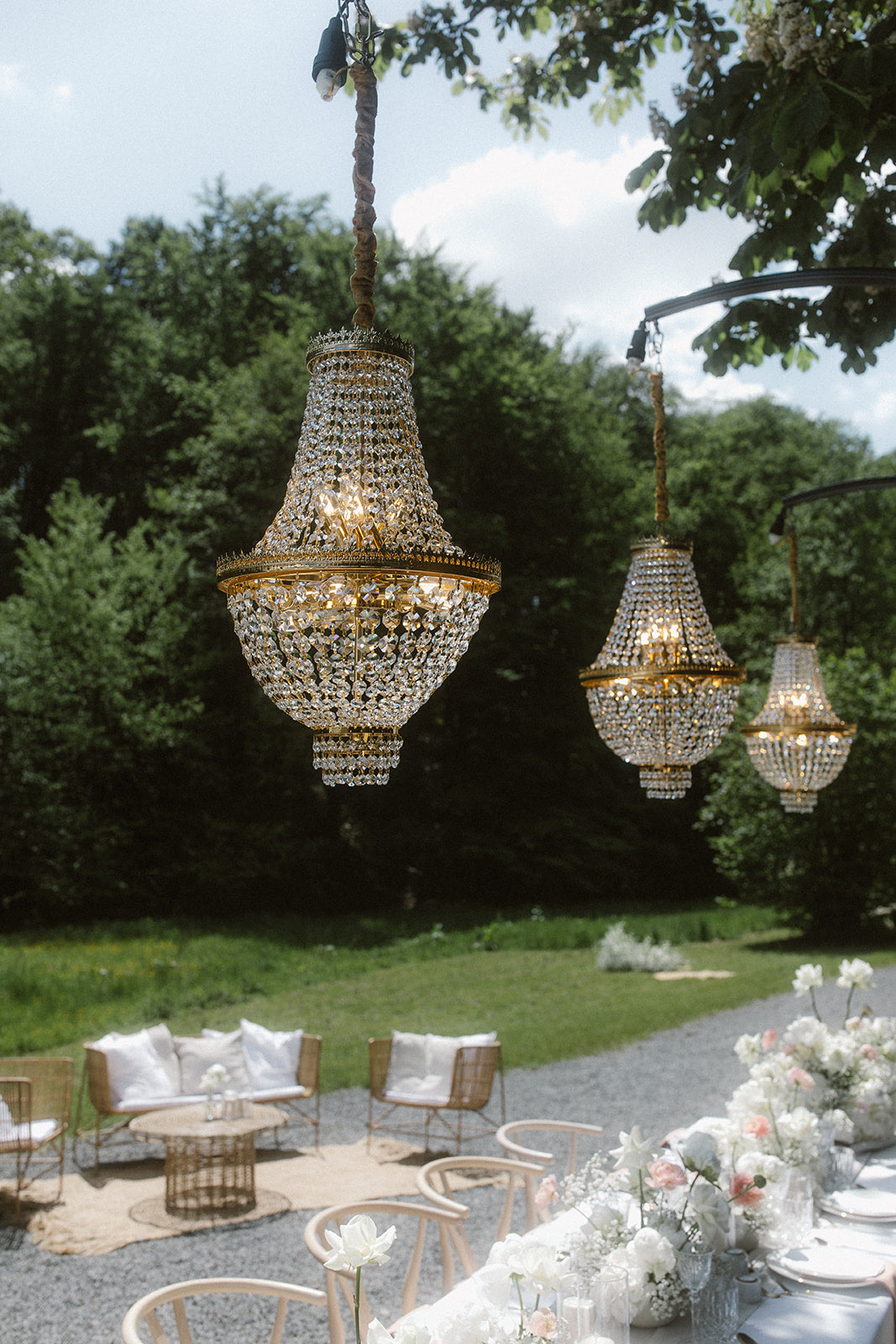 Hängende, ästhetische Kronleuchter bei einer eleganten Outdoor-Hochzeit mit gemütlicher Lounge-Ecke im Hintergrund. Diese luxuriöse Outdoor-Hochzeitsdekoration kombiniert funkelnde Kronleuchter mit einer einladenden Lounge-Ecke im Garten. Die harmonische Mischung aus Eleganz und Komfort schafft eine festliche und zugleich entspannte Atmosphäre für die Gäste.