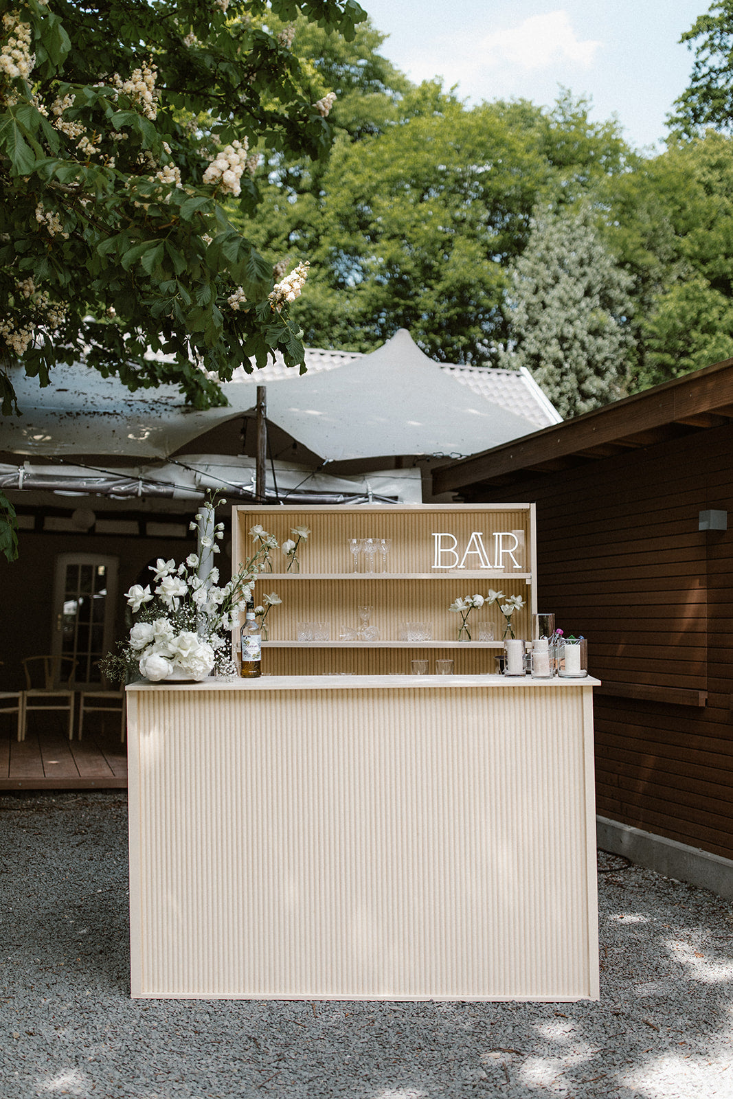 Outdoor-Hochzeitsbar mit minimalistischer Blumendekoration und Kristallgläsern im Hintergrund. Diese schlichte Outdoor-Hochzeitsbar ist mit eleganter Blumendekoration und feinen Kristallgläsern ausgestattet. Das natürliche, minimalistische Design fügt sich perfekt in die Umgebung ein und bietet eine stilvolle Anlaufstelle für die Gäste während der Feier.