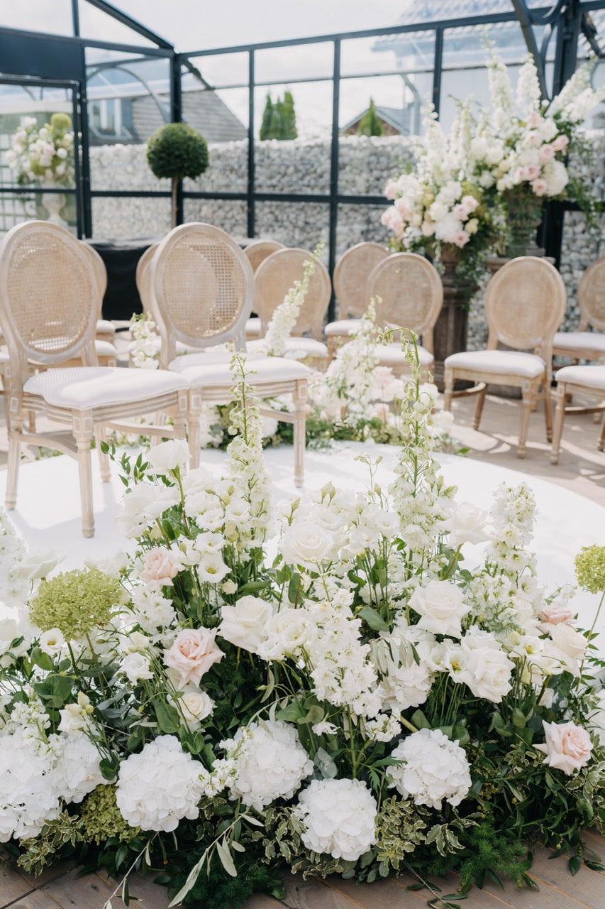 Ästhetisches Hochzeitskonzept mit einem eleganten Zeremonie Bereich mit weißen und rosafarbenen Blumen vor eleganten Stühlen in einer Orangerie. Dieser wunderschöne Zeremonie Bereich ist mit üppigen weißen und rosafarbenen Blumenarrangements gestaltet, die den Gang und die eleganten Stühle schmücken. Die Glaswände der Orangerie sorgen für viel Tageslicht und verleihen der Zeremonie eine romantische, natürliche Atmosphäre. Perfekt für eine elegante und stilvolle Hochzeitsfeier.