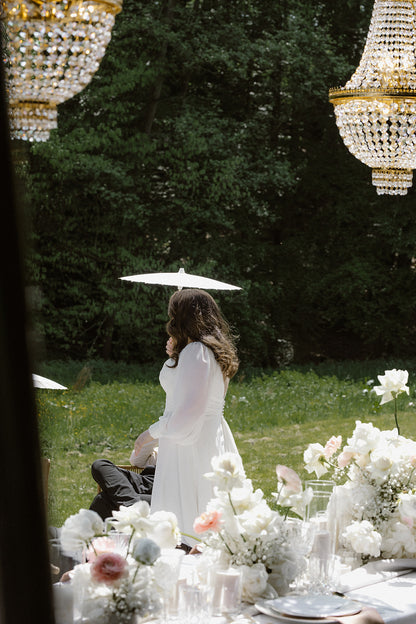 Braut im Garten mit weißem Papierschirm, umgeben von eleganter Hochzeitsdekoration und Kronleuchtern in Köln. Diese romantische Szene zeigt eine Braut, die im Garten mit einem weißen Papierschirm flaniert. Im Vordergrund sind edle Hochzeitsdekorationen mit weißen Blumen und funkelnden Kronleuchtern zu sehen, was eine stilvolle und verträumte Atmosphäre schafft.