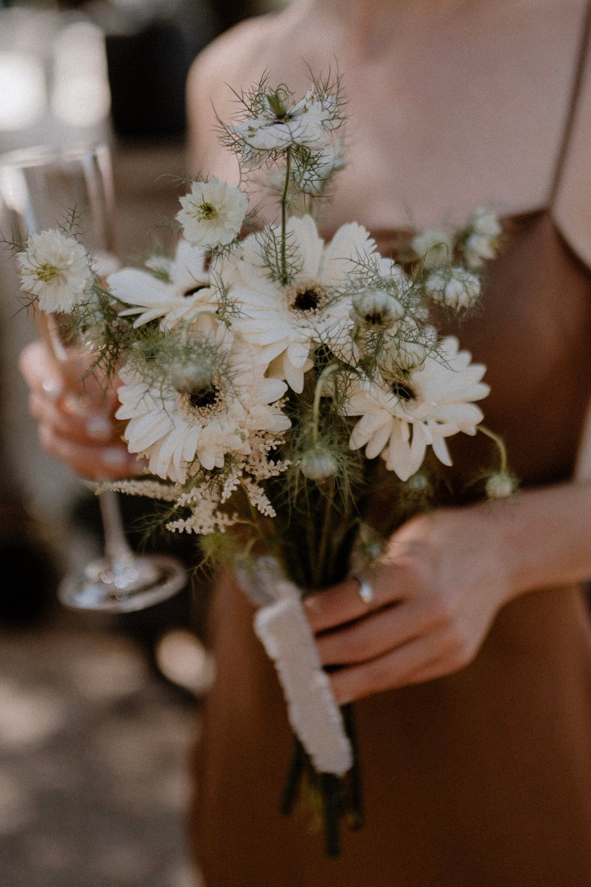 Brautjungfer mit schlichtem Brautstrauß aus weißen Blumen und einem Sektglas in der Hand in Hamburg. Schlichter Brautstrauß aus weißen Blumen, gehalten von einer Brautjungfer in einem dezenten Kleid, ideal für eine zeitlose und charmante Hochzeitsfeier im Freien.