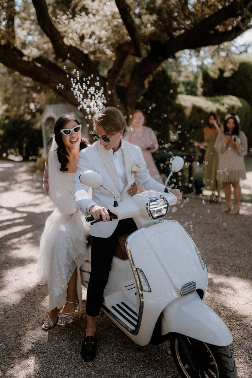 Brautpaar auf einer weißen Vespa, umgeben von Seifenblasen bei einer Hochzeit im Freien deutschlandweit. Das Brautpaar fährt stilvoll auf einer weißen Vespa, umgeben von Seifenblasen, während Freunde sie bei einer charmanten Hochzeitsfeier im Freien feiern.