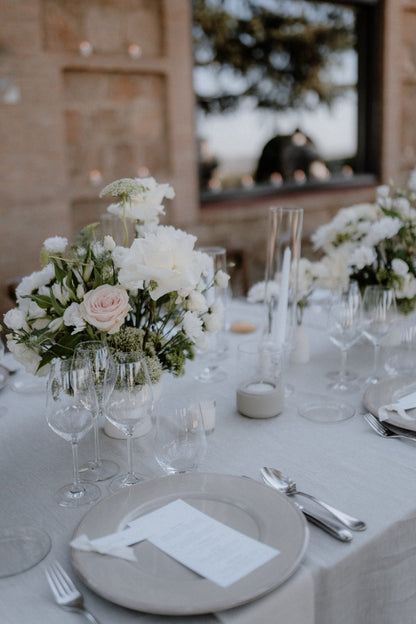 Cleane Hochzeitsdekoration mit weißen Blumen und hohen Kerzen. Ein elegant gedeckter Hochzeitstisch mit weißen Blumenarrangements, hohen Kerzen und schlichten Gläsern, ideal für eine zeitlose und charmante Hochzeitsfeier im Freien.
