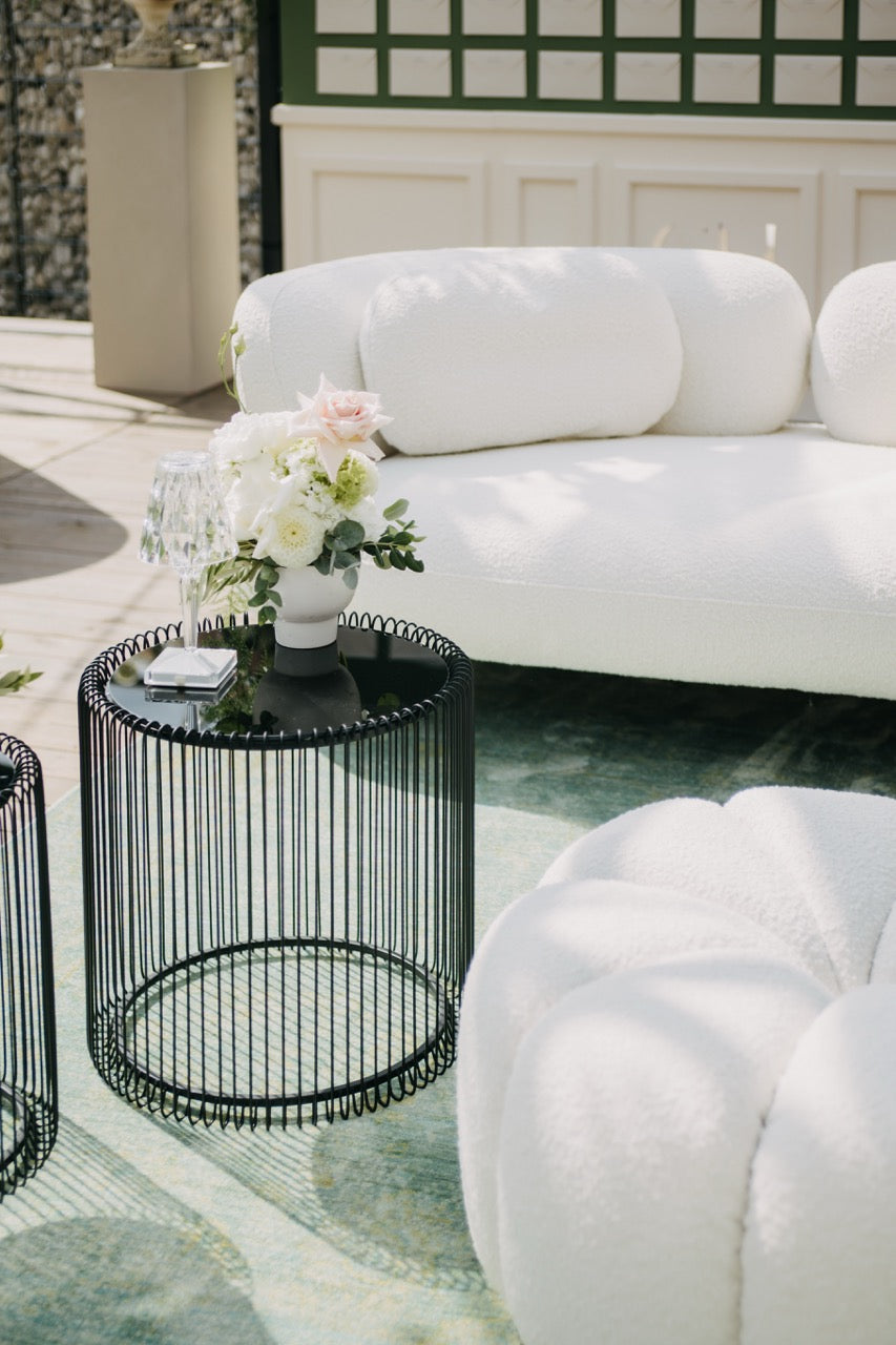 Moderner Lounge-Bereich mit weißen Polstermöbeln und einem minimalistischen Blumenarrangement auf einem stilvollen Beistelltisch. Dieser elegante Lounge-Bereich kombiniert moderne weiße Polstermöbel mit einem schwarzen, filigranen Beistelltisch. Ein dezentes Blumenarrangement verleiht dem Setting einen Hauch von Romantik und vervollständigt die stilvolle Atmosphäre. Perfekt für eine Hochzeit, die modernen Chic mit Eleganz verbindet.