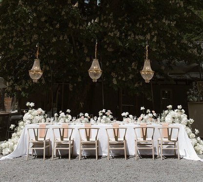 Edle Hochzeitsdekoration mit langem Tisch, weißen Blumenarrangements und hängenden Kronleuchtern im Freien. Diese romantische und elegante Hochzeitsdekoration zeigt einen langen, festlich gedeckten Tisch im Freien, geschmückt mit üppigen weißen Blumenarrangements und stilvollen, hängenden Kronleuchtern. Ideal für eine Outdoor-Hochzeit mit luxuriösem Flair.