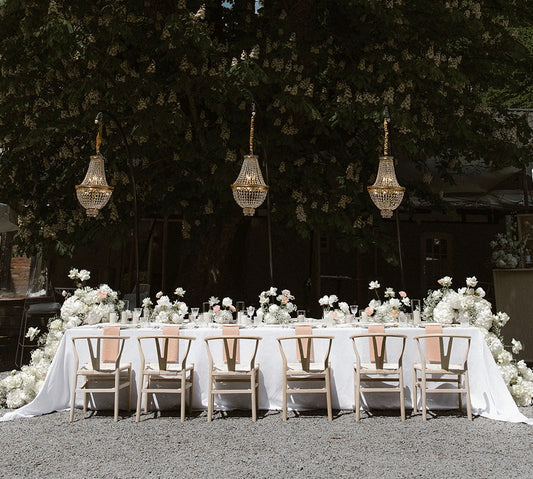 Edle Hochzeitsdekoration mit langem Tisch, weißen Blumenarrangements und hängenden Kronleuchtern im Freien. Diese romantische und elegante Hochzeitsdekoration zeigt einen langen, festlich gedeckten Tisch im Freien, geschmückt mit üppigen weißen Blumenarrangements und stilvollen, hängenden Kronleuchtern. Ideal für eine Outdoor-Hochzeit mit luxuriösem Flair.