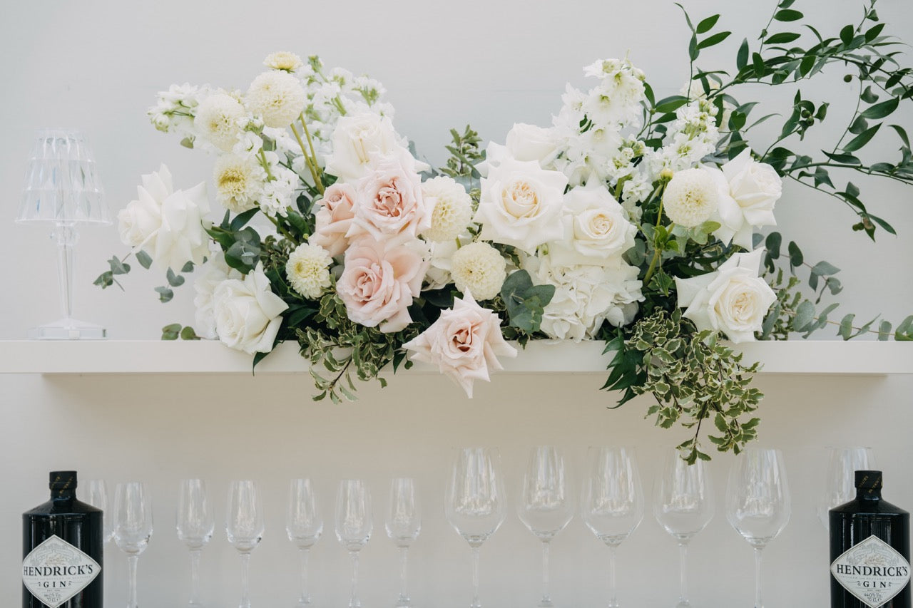 Elegante Blumendekoration mit weißen und rosafarbenen Rosen über einer stilvollen Bar mit Gläsern und Flaschen. Diese geschmackvolle Blumendekoration aus weißen und rosafarbenen Rosen schmückt die Bar und verleiht ihr eine romantische, edle Note. Die klaren Gläser und stilvoll präsentierten Flaschen unterstreichen das luxuriöse Ambiente und schaffen eine ansprechende Kulisse für einen eleganten Empfangsbereich bei einer Hochzeit.