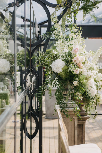 Ein prächtiges Blumenarrangement mit weißen und rosafarbenen Blüten in einer Orangerie in Berlin. Dieses große Blumenarrangement aus weißen und rosafarbenen Blüten, kombiniert mit üppigem Grün, schmückt stilvoll die Seiten der Orangerie. Die reflektierende Glasstruktur und die floralen Details schaffen eine harmonische Verbindung zwischen Natur und moderner Eleganz. Ideal für eine exklusive Hochzeit mit romantischer Atmosphäre.