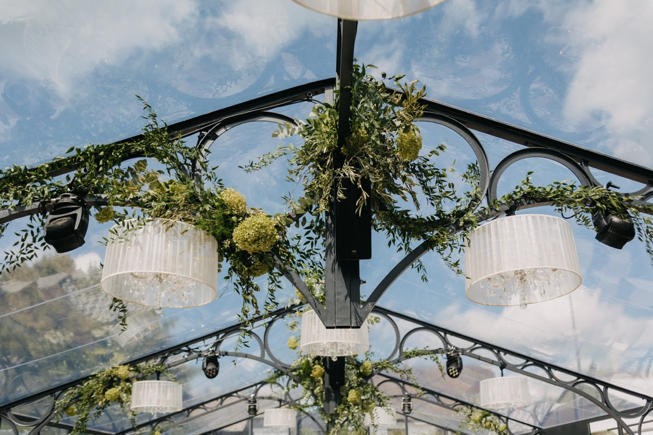 Dekorierte Decke einer Orangerie mit eleganten Kronleuchtern und grünen Blumengirlanden. Die Decke der Orangerie ist mit stilvollen Kronleuchtern und üppigen Blumengirlanden geschmückt, die dem Raum eine romantische und luxuriöse Atmosphäre verleihen. Die Kombination aus Glasdach und floralen Details lässt natürliches Licht herein und schafft eine elegante Kulisse für eine exklusive Hochzeitsfeier.