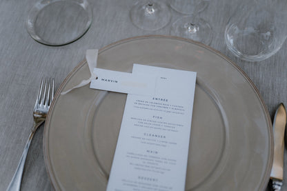 Schlichtes und ästhetisches Hochzeitsgedeck mit nude Platzteller, Menükarte und Namenskarte. Elegantes Hochzeitsgedeck mit einer schlichten Menükarte und einer dezenten Namenskarte auf einem nudefarbenen Platzteller, perfekt für eine minimalistische und zeitlose Hochzeitsdekoration.