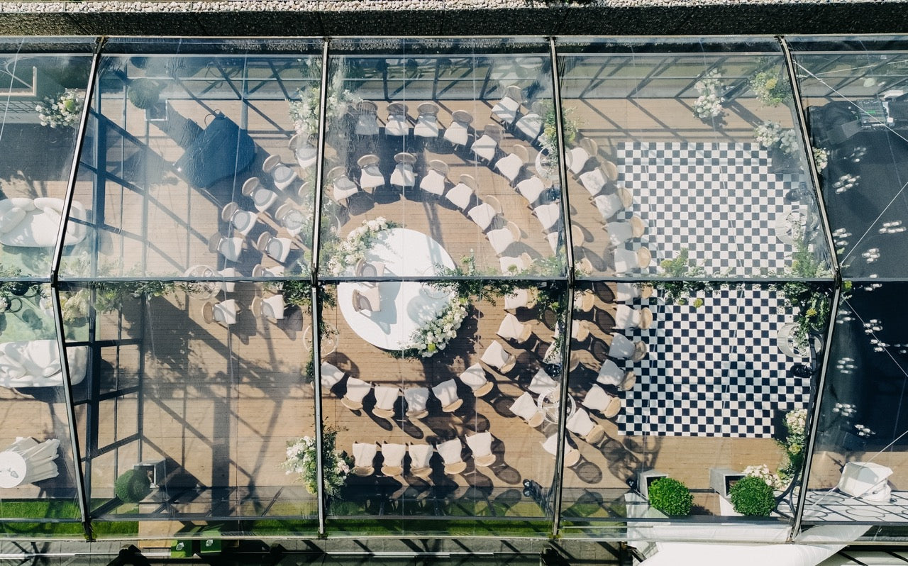 Orangerie als Hochzeitslocation mit rundem Trausetting und Schachbrett-Tanzfläche, aus der Vogelperspektive aufgenommen. Die elegante Hochzeitslocation zeigt ein perfekt arrangiertes Trausetting und eine stilvolle Schachbrett-Tanzfläche. Die Struktur der Orangerie lässt viel Tageslicht hinein, was eine harmonische und luxuriöse Atmosphäre schafft. Ideal für Paare, die eine moderne und stilvolle Hochzeitsfeier planen.