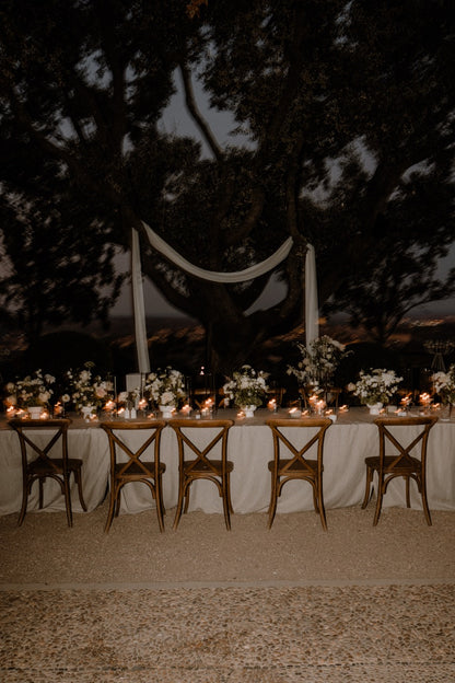 Hochzeitstafel im Freien bei Nacht, dekoriert mit Lichtern und weißen Blumengestecken. Stimmungsvolle Abenddekoration einer Hochzeitstafel im Freien, beleuchtet mit Kerzen und Lichtern, verziert mit weißen Blumen, ideal für eine romantische und zeitlose Feier unter freiem Himmel.