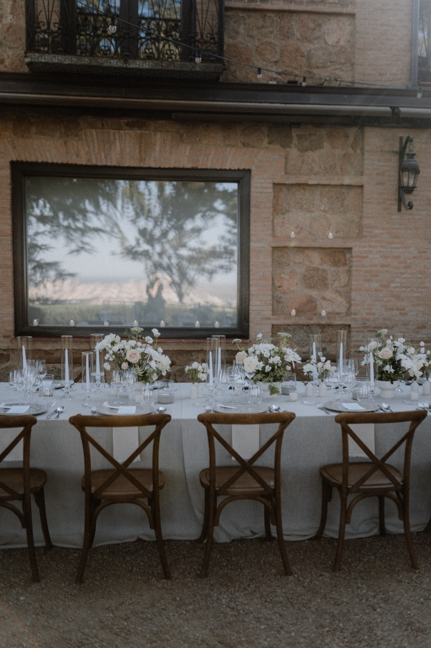 Hochzeitstafel im Freien mit Crossback Stühlen und weißen Blumen vor einem steinernen Gebäude.
Eine schlichte Hochzeitstafel im Freien, dekoriert mit weißen Blumenarrangements und rustikalen Crossback Stühlen, ideal für eine charmante und zeitlose Feier im Freien.