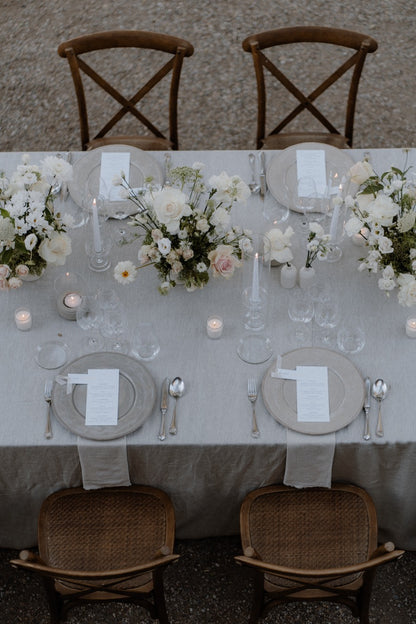 Schlicht gedeckter Hochzeitstisch mit weißen Blumenarrangements und rustikalen Holzstühlen. Eleganter Hochzeitstisch im Freien mit weißen Blumenarrangements, dezenten Kerzen und rustikalen Holzstühlen, ideal für eine minimalistische und zeitlose Feier.