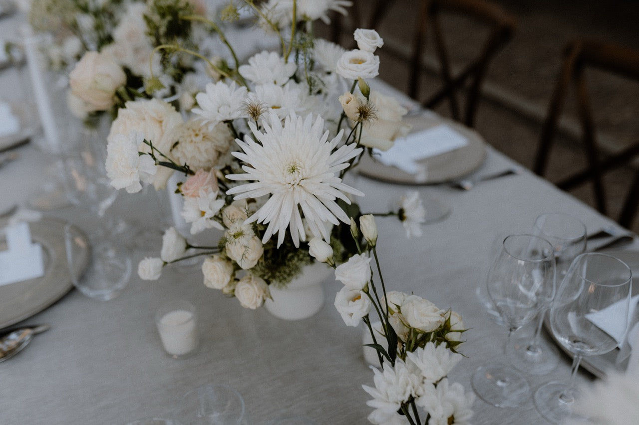 Schlichte und elegante Hochzeitstischdekoration mit weißen Blumen und dezenten Gläsern. Stilvolle Hochzeitstischdekoration mit weißen Blumenarrangements und zurückhaltendem Geschirr, ideal für eine zeitlose und charmante Hochzeitsfeier.