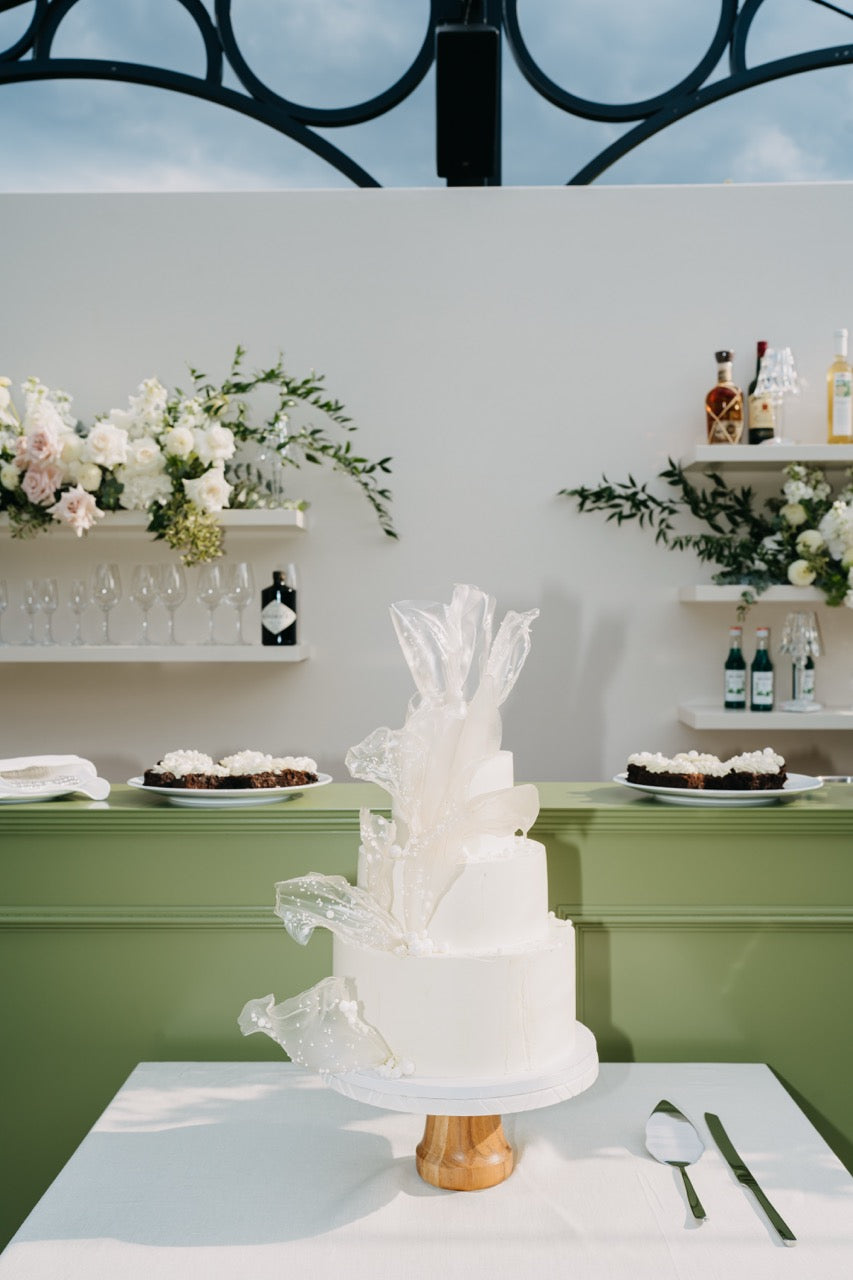 Minimalistische, elegante Hochzeitstorte mit weißen Zuckerdekorationen vor einer stilvollen Bar mit Blumenarrangements. Die dreistöckige, schneeweiße Hochzeitstorte besticht durch ihre minimalistische Eleganz und kunstvolle Zuckerdekorationen. Im Hintergrund runden dezente Blumenarrangements und eine stilvoll eingerichtete Bar das luxuriöse Gesamtbild ab, was eine perfekte Mischung aus Modernität und Raffinesse für eine gehobene Hochzeitsfeier schafft.