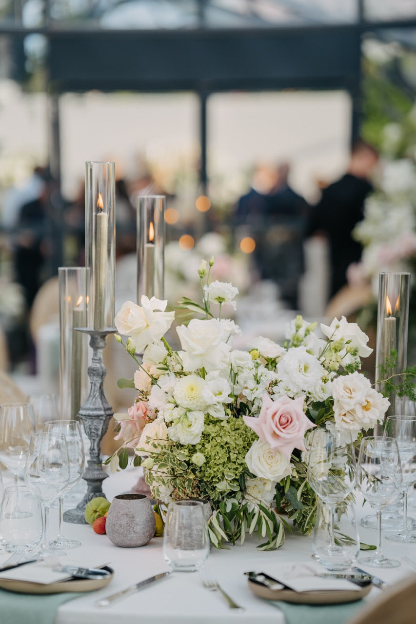 Elegantes Blumenarrangement mit weißen und rosafarbenen Blüten sowie hohen Kerzen auf einem festlich gedeckten Hochzeitstisch. Das luxuriöse Blumenarrangement mit weißen Rosen, zarten Rosa-Akzenten und grünen Blättern ergänzt perfekt die eleganten, hohen Kerzenständer. Dieser opulent gedeckte Hochzeitstisch strahlt Exklusivität und Raffinesse aus und ist ideal für eine stilvolle und hochwertige Hochzeitsfeier.