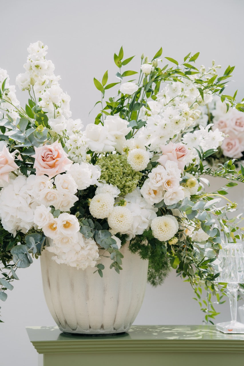 Üppiges Blumengesteck mit weißen und rosafarbenen Blumen in einer eleganten Vase bei einer luxuriösen Hochzeit. Dieses prachtvolle Blumengesteck aus weißen Rosen und zarten rosafarbenen Blüten, kombiniert mit grünen Akzenten, verleiht der Hochzeitsdekoration eine romantische und luxuriöse Note. Arrangiert in einer eleganten weißen Vase, ist es ein wahrer Blickfang und unterstreicht den exklusiven Stil der Feier. Perfekt für eine stilvolle Hochzeitsdekoration, die klassische Eleganz ausstrahlt.