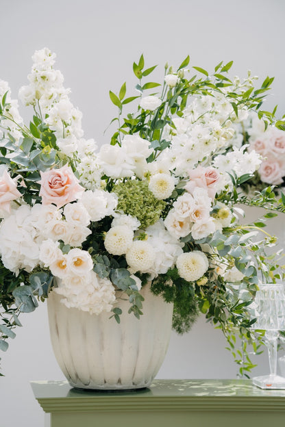 Üppiges Blumengesteck mit weißen und rosafarbenen Blumen in einer eleganten Vase bei einer luxuriösen Hochzeit. Dieses prachtvolle Blumengesteck aus weißen Rosen und zarten rosafarbenen Blüten, kombiniert mit grünen Akzenten, verleiht der Hochzeitsdekoration eine romantische und luxuriöse Note. Arrangiert in einer eleganten weißen Vase, ist es ein wahrer Blickfang und unterstreicht den exklusiven Stil der Feier. Perfekt für eine stilvolle Hochzeitsdekoration, die klassische Eleganz ausstrahlt.