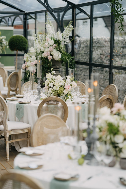 Elegante Tischdekoration mit weißen Blumen und hohen Kerzen in einem luxuriösen Glashaus bei einer Hochzeitsfeier deutschlandweit. Die festlichen Tische, geschmückt mit üppigen weißen Blumenarrangements und hohen, eleganten Kerzen, verleihen der Hochzeit im stilvollen Glashaus eine exklusive und romantische Atmosphäre. Die sanften Farbtöne und edlen Materialien betonen den luxuriösen Charakter dieser außergewöhnlichen Hochzeitsdekoration.