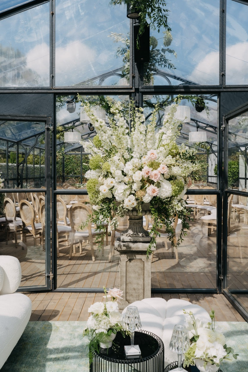 Großes, elegantes Blumengesteck mit weißen und rosafarbenen Blumen in einer stilvollen Orangerie bei einer luxuriösen Hochzeit. Dieses prächtige Blumengesteck aus weißen und rosafarbenen Blüten steht auf einem eleganten Sockel und schmückt den Eingang einer luxuriösen Orangerie. Die harmonische Kombination aus üppigen Blüten und modernem Glashaus-Design schafft eine atemberaubende Kulisse für eine exklusive Hochzeitsfeier.