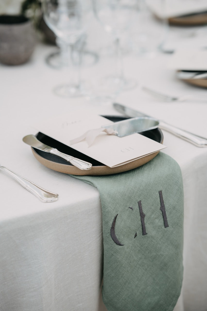 Ästhetische, personalisierte Serviette mit Monogramm und elegantes Tischgedeck bei einer luxuriösen Hochzeit. Dieses exklusive Tischgedeck besticht durch eine personalisierte Serviette mit Monogramm und edlem Besteck auf einem minimalistischen Teller. Die schlichte, elegante Tischdekoration fügt sich harmonisch in das luxuriöse Ambiente der Hochzeit ein und verleiht der Tafel eine individuelle Note. Perfekt für Paare, die Liebe zum Detail und Stil schätzen.