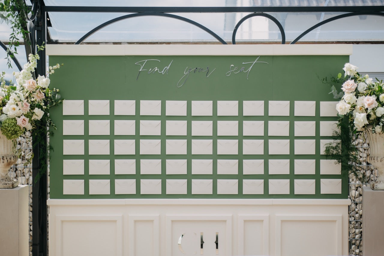 Elegante und ästhetische, grüne Sitzplanwand mit weißen Umschlägen, umrahmt von Blumendekoration. Dieser stilvolle Sitzplan besticht durch seine schlichte grüne Wand mit perfekt angeordneten weißen Platzkarten. Umrahmt von üppigen Blumenarrangements in sanften Farbtönen, fügt sich das Display harmonisch in die luxuriöse Hochzeitsdekoration ein. Eine elegante Lösung, um den Gästen ihre Sitzplätze stilvoll und übersichtlich zu präsentieren.