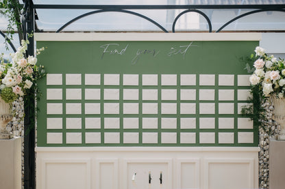 Elegante und ästhetische, grüne Sitzplanwand mit weißen Umschlägen, umrahmt von Blumendekoration. Dieser stilvolle Sitzplan besticht durch seine schlichte grüne Wand mit perfekt angeordneten weißen Platzkarten. Umrahmt von üppigen Blumenarrangements in sanften Farbtönen, fügt sich das Display harmonisch in die luxuriöse Hochzeitsdekoration ein. Eine elegante Lösung, um den Gästen ihre Sitzplätze stilvoll und übersichtlich zu präsentieren.