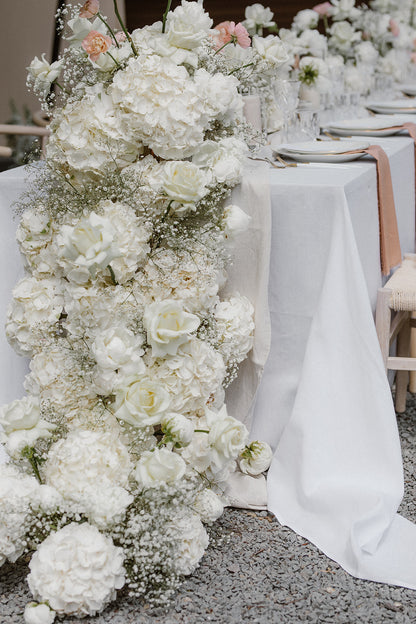 Üppige weiße Blumendekoration aus Hortensien entlang eines Hochzeitstisches. Diese opulente Hochzeitsdekoration besticht durch eine üppige Blumenarrangement aus weißen Hortensien und Schleierkraut, das entlang eines Hochzeitstisches drapiert ist. Perfekt für eine elegante und romantische Hochzeitsfeier mit luxuriösem Blumenkonzept.
