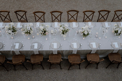 Schlichte Hochzeitstafel im Freien in Hamburg mit weißen Tischdecken, Blumenarrangements und Holzstühlen. Eine elegante, lange Hochzeitstafel im Freien, dekoriert mit weißen Tischdecken, dezenten Blumenarrangements und rustikalen Holzstühlen. Perfekt für eine zeitlose und charmante Hochzeitsfeier.
