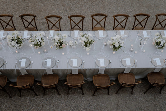 Schlichte Hochzeitstafel im Freien in Hamburg mit weißen Tischdecken, Blumenarrangements und Holzstühlen. Eine elegante, lange Hochzeitstafel im Freien, dekoriert mit weißen Tischdecken, dezenten Blumenarrangements und rustikalen Holzstühlen. Perfekt für eine zeitlose und charmante Hochzeitsfeier.