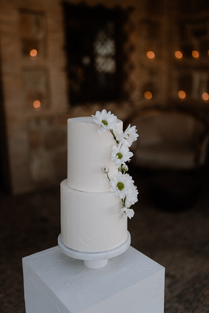 Schlichte und ästhetische zweistöckige Hochzeitstorte mit weißen Blumen dekoriert. Minimalistische, zweistöckige Hochzeitstorte, verziert mit weißen Blumen, perfekt für eine elegante und zeitlose Hochzeitsfeier.