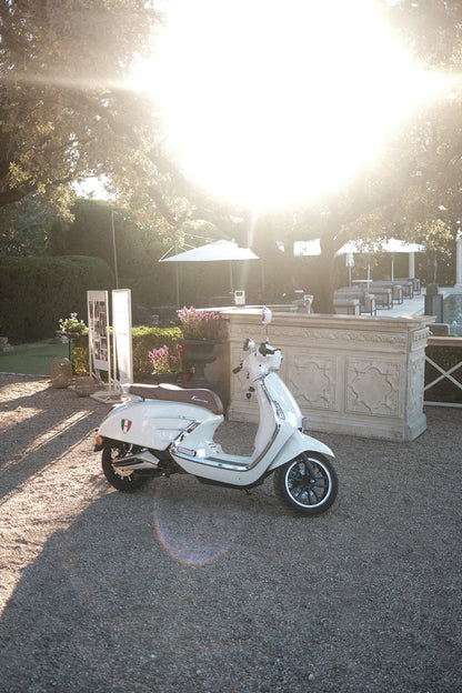 Weiße Vespa als Hochzeitsdekoration im Freien bei Sonnenuntergang. Stilvolle weiße Vespa als dekoratives Element auf einer charmanten Hochzeitsfeier im Freien, beleuchtet vom warmen Licht des Sonnenuntergangs, ideal für eine romantische Atmosphäre.